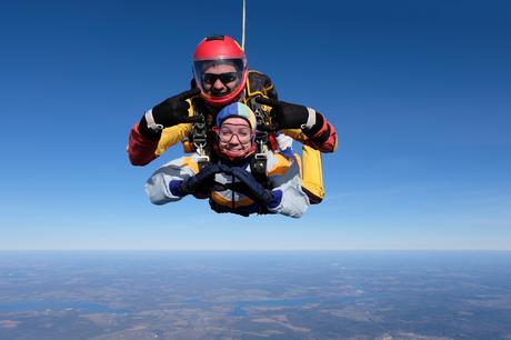 Saut en parachute en tandem en plein ciel, avec un instructeur et une personne équipée de combinaisons colorées et de harnais de sécurité. Expression de joie et d’excitation sur le visage du parachutiste, bras formant un symbole avec les mains. En arrière-plan, un vaste paysage avec l’horizon, un ciel bleu dégagé et une vue panoramique sur la terre en contrebas. Expérience d’adrénaline et de liberté en altitude.