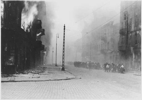 Photo d'une rue du ghetto. A gauche un immeuble brûle, des flammes sortent du premier étage. Sur la droite de la rue, assez loin, des groupes de civils marchent dans la fumée en s'éloignant de l'objectif.