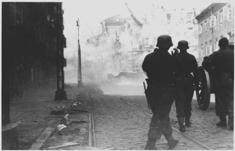 Photo très contrastée : au fond, très clair, immeuble en cours de destuction avec de la fumée. Au premier plan, très sombre, 3 silhouettes de soldats allemands autour d'un canon.