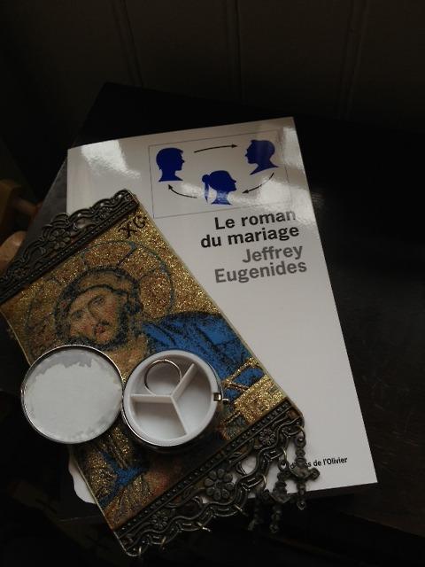 Si Jeffrey Eugenides avait écrit plus tôt dans le temps, il serait probablement apparu dans la liste de Neil Hannon (cf chanson précédente).
(Mais, au moment où Promenade était enregistré, il n’avait publié qu’un livre, certes remarqué (Virgin Suicides) que pour ma part je n’ai pas -encore- lu.)
Bref, peu importe, (il a écrit trois livres en trente ans, dont Middlesex -Prix Pullitzer de la Fiction), je voulais seulement partager mon émotion face à son dernier roman, Le Roman du Mariage, publié cette année. Pour commencer, j’ai furtivement réalisé une photographie où j’ai gracieusement mis en scène les éléments de l’histoire, devinant que ce serait difficile d’en parler.
Je crois que nous avons là un grand livre. Pourquoi ? C’est ce qui est difficile à dire. J’ai lu diverses critiques deci-delà, toutes élogieuses (quoique peu nombreuses ai-je trouvé compte-tenu de l’événement littéraire à mon avis). Apparemment, l’auteur se serait demandé comment écrire un roman sur le mariage après que le mariage ne constitue plus l’affaire de toute une vie, comme il l’était dans les romans de Jane Austen par exemple. D’ailleurs, il y a un point commun entre Jane Austen et lui, malgré la modernité de son écriture et le fait que l’intrigue se situe aux USA au début des années 80 : comme chez la célèbre britannique, la psychologie des personnages est tellement fouillée et précise, que le moindre fait narratif dans les interactions du trio amoureux (prendre un thé ou boire un verre avec telle personne) prend lors de la lecture l’ampleur des rebondissements d’un thriller. C’est sans doute cela que j’ai particulièrement adoré. Entre Madeleine, Mitchell et Leonard, les sentiments circulent, et ça nous passionne, sans aucune mièvrerie, alors qu’il s’agit d’amour, sujet qui peut vite devenir glissant et tellement convenu. 
Ensuite, ce que j’ai aimé, ce sont les “expériences” auxquelles se livrent les trois protagonistes et qui les transmutent de gré ou de force dans l’âge adulte. L’histoire débute au moment où ils terminent leur cursus universitaire.
Madeleine nous est présentée dès la première page par le biais de sa bibliothèque personnelle, (elle qui rédigeait un mémoire sur le roman du mariage, sur un corpus allant d’Austen à Updike). Elle est une booklover elle aussi, complètement pénétrée de cette passion, immergée dans ce monde ; voilà pourquoi la présenter à travers ce prisme est particulièrement pertinent (et cela m’a aussi rappelé les douces heures de la fac de Lettres…). C’est un personnage extrêmement attachant, même si principalement évoqué à travers des prismes -en même temps, c’est sans doute la meilleure façon d’évoquer son caractère doux, loyal et empathique-, celui de son amour des livres, puis celui de son milieu social, puis celui de son difficile rôle auprès de celui qu’elle aime.
Mitchell, esprit brillant mais trop sage pour vraiment jouir de ses années de fac sur le plan sexuel ou sentimental, est d’une honnêteté bouleversante. Il essaie d’être cohérent face à ses interrogations nombreuses et sa soif de spiritualité. Éconduit par Madeleine, il part en voyage sur les autres continents, jusqu’à aller au bout de sa logique et se retrouver à panser des plaies chez Mère Teresa à Calcutta.
Leonard, apparaît au départ comme une sorte de garçon idéal : excessivement intelligent, beau, vif, drôle, incisif, intense, ayant la capacité de rendre le réel plus grand, de vous enrober de son attention en vous surélevant le temps de son écoute. Hélas, toutes ces capacités qui font de lui un être exceptionnel, sont la face positive de ce qui fait de lui littéralement un être extraordinaire : la maniaco-dépression. Il s’enfoncera ensuite dans l’autre versant de la maladie, subissant ensuite différentes phases : manie, dépression, soins, médicamentation, lutte contre les effets secondaires, auto-dosage, rechutes, etc… Eugenides nous plonge au coeur de ce trouble, des montagnes russes de son esprit, c’est d’une précision extrême, très détaillé et passionnant. On voit Leonard lutter contre lui-même, son enfance, la chimie de son cerveau, c’est bouleversant. Rarement un personnage ne m’a autant émue.
Alors, le schéma traditionnel et rebattu Mitchell-qui-aime-Madeleine-qui-aime-Leonard échappe au conventionnel car l’écriture d’Eugenides lie leurs trois destins avec une précision clinique, une ironie discrète quant aux illusions de la jeunesse -notamment la lettrée, celle qu’il a connue-, et aux modes littéraires des universités et un sens du dramatique qui ne semble jamais forcé. Son écriture est à la fois simple, dévouée toute entière à la justesse des émotions et des idées, et pleine de surprises. J’ai parfois souri en lisant des comparaisons neuves, et pourtant parfaitement parlantes. Presque six cents pages, et aucune de trop. 
Bref, il a réussi, en 2013, à faire lui aussi son roman sur le mariage ; en effet, choisir son époux reste un enjeu de taille pour réussir sa vie, même si l’erreur est moins coûteuse qu’au dix-neuvième siècle. Madeleine n’est pas un personnage tragique comme Isabel Archer de James. Le véritable enjeu se déplace légèrement et aimer quelqu’un devient une métaphore de devenir adulte, pas juste éprouver des sentiments vibrants pour quelqu’un, ce que l’on fait aisément dans sa jeunesse, mais être auprès de quelqu’un, tous les jours, avec tous les détails peu glamours. Peut-être est-ce cela qui est touchant aussi : Madeleine aime, souffre, se dévoue, échoue partiellement, mais ce n’est pas tragique, c’est dénué de cette grandeur habituellement accordée aux héros de fiction. C’est juste triste, dur, mais banal, un accident de parcours qui se corrigera, on le devine. Madeleine, l’héroïne d’un livre de 2013, dont l’héroïsme est condamné à rester modeste.
Je réalise que j’ai écrit une tartine sans bien transmettre ce que je voulais. Je me tais maintenant, j’en reparlerai peut-être tant je suis saisie.