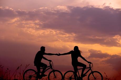 Lovers share a silhouetted bicycle ride, hands reaching out to each other