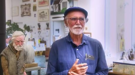 Joe Fafard se tient devant la caméra et porte un chapeau avec une chemise bleue. On voit derrière lui de nombreuses œuvres d'art.